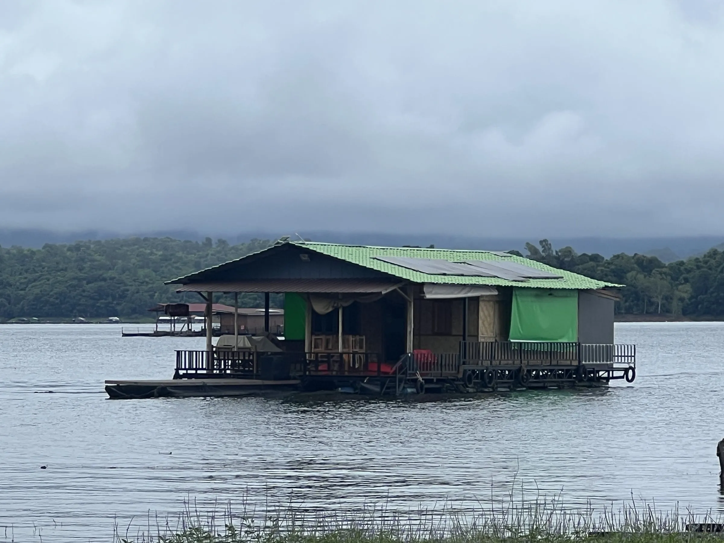 A green-roofed wooden raft house moored on the still water of Khao Laem Lake with forested hills behind.