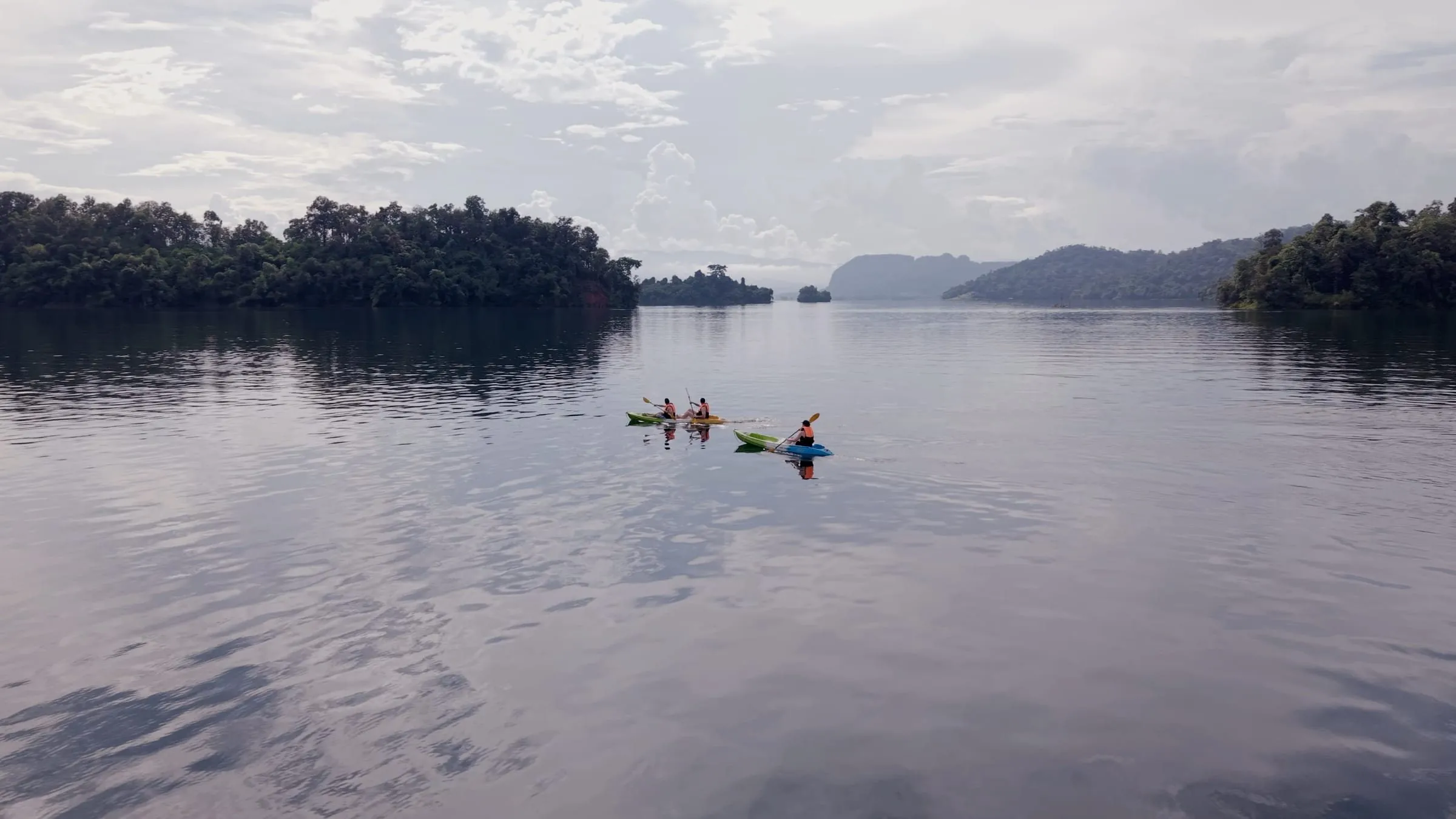 Two kayakers out on the open grey water of Khao Laem Lake, forested headlands beyond.